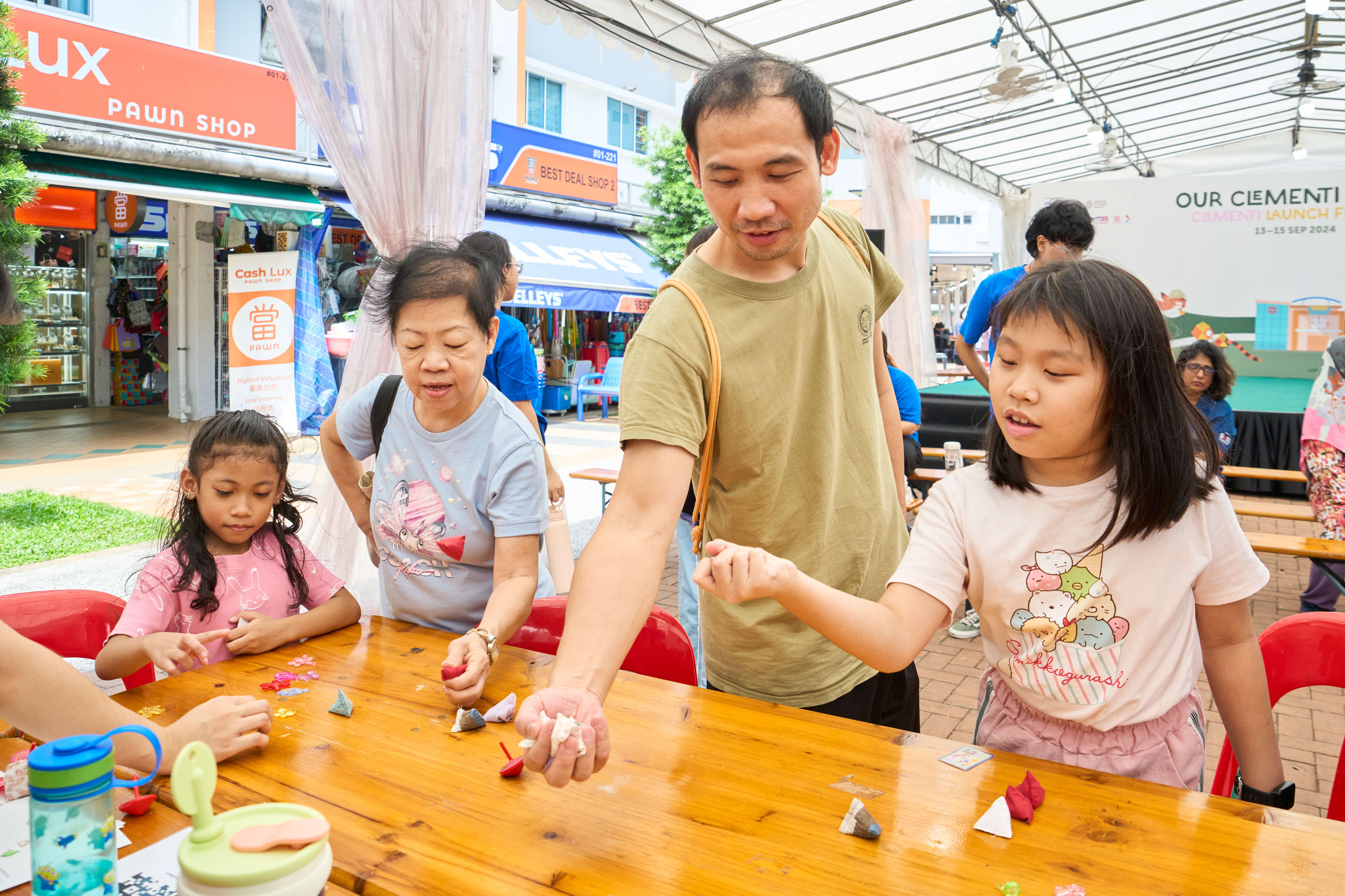A group of people are gathered around a wooden table doing crafts at an outdoor community event. There are shops in the background.